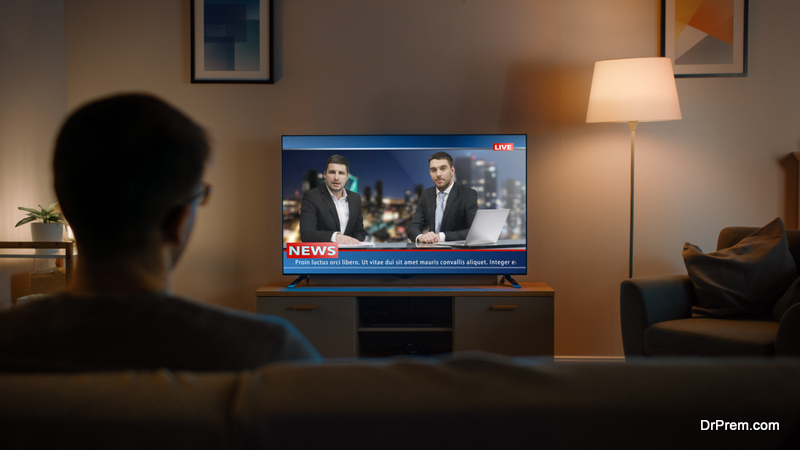Young Man in Glasses is Sitting on a Sofa and Watching TV with Live News