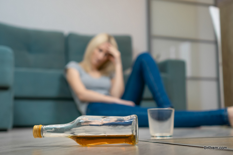 Young blonde woman is sitting on the floor near the sofa and holding her head, in the foreground is a half-empty bottle of whiskey and a glass